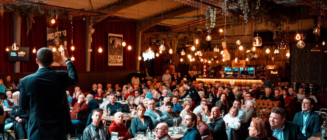 A packed event room seen from behind a speaker, every table taken by seated guests under rows of warm filament bulbs.